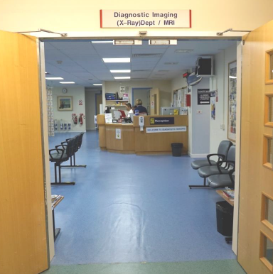 The entrance to a hospital department. Through the door you can see a reception desk and a waiting area. Above the door there is a sign that reads: "Diagnostic Imaging (X-Ray) Department / MRI".