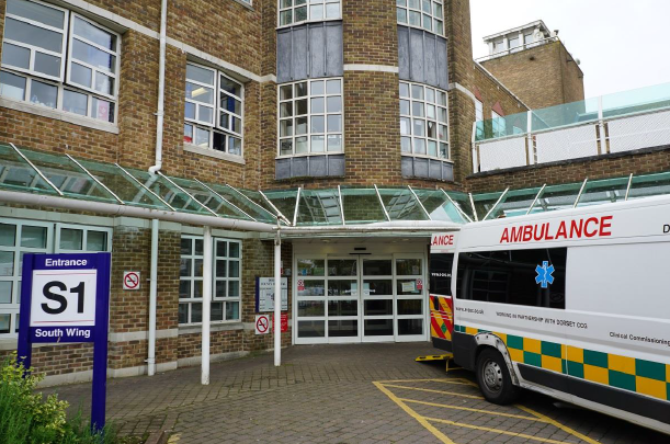 The outside entrance of a hospital building. There is an ambulance parked outside with its doors open and a sign that reads: "Entrance S1 South Wing".