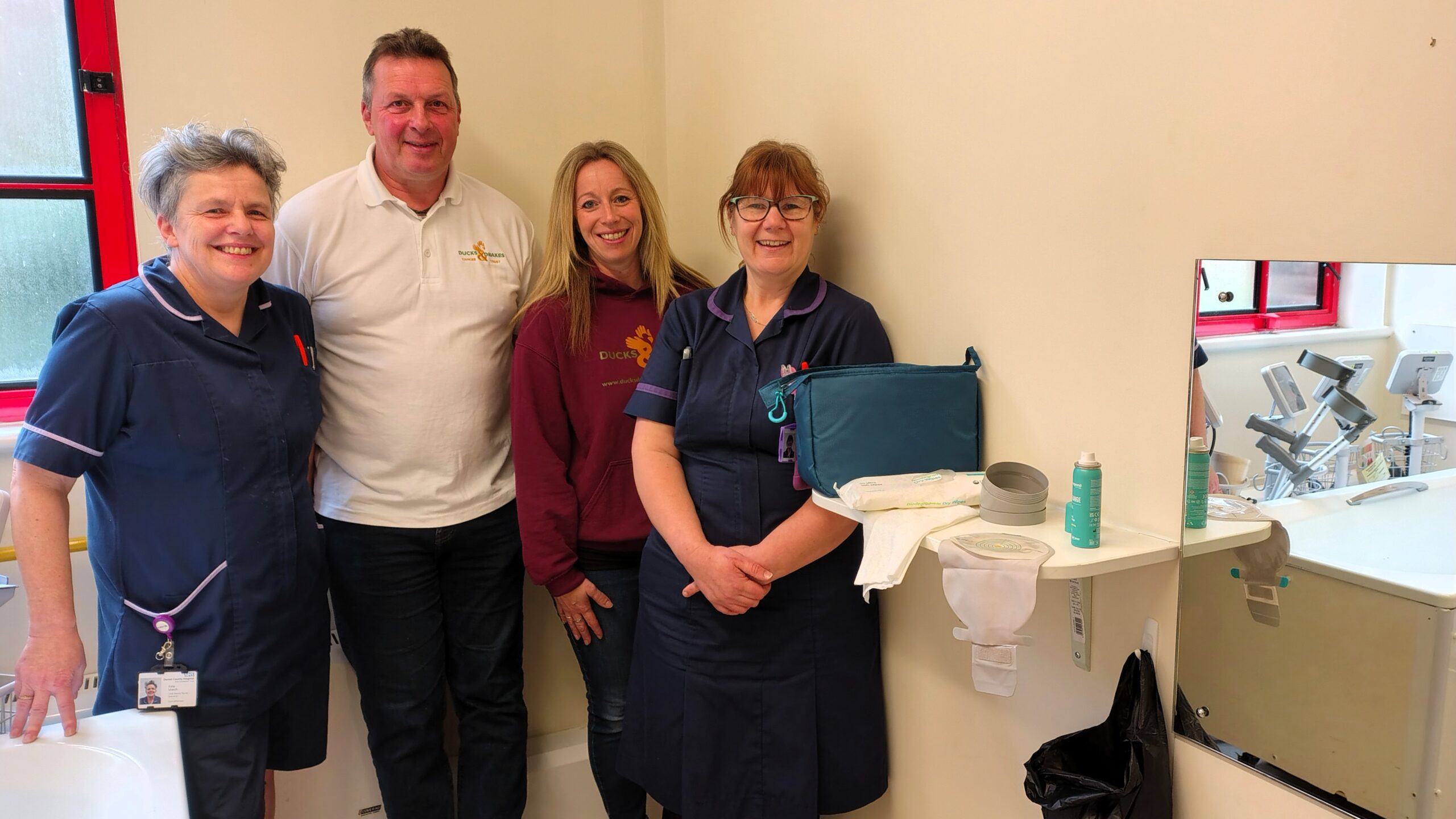 A group of four people stand smiling in a room with medical equipment. Two of them are wearing uniforms. The room includes a sink and a wall-mounted mirror.