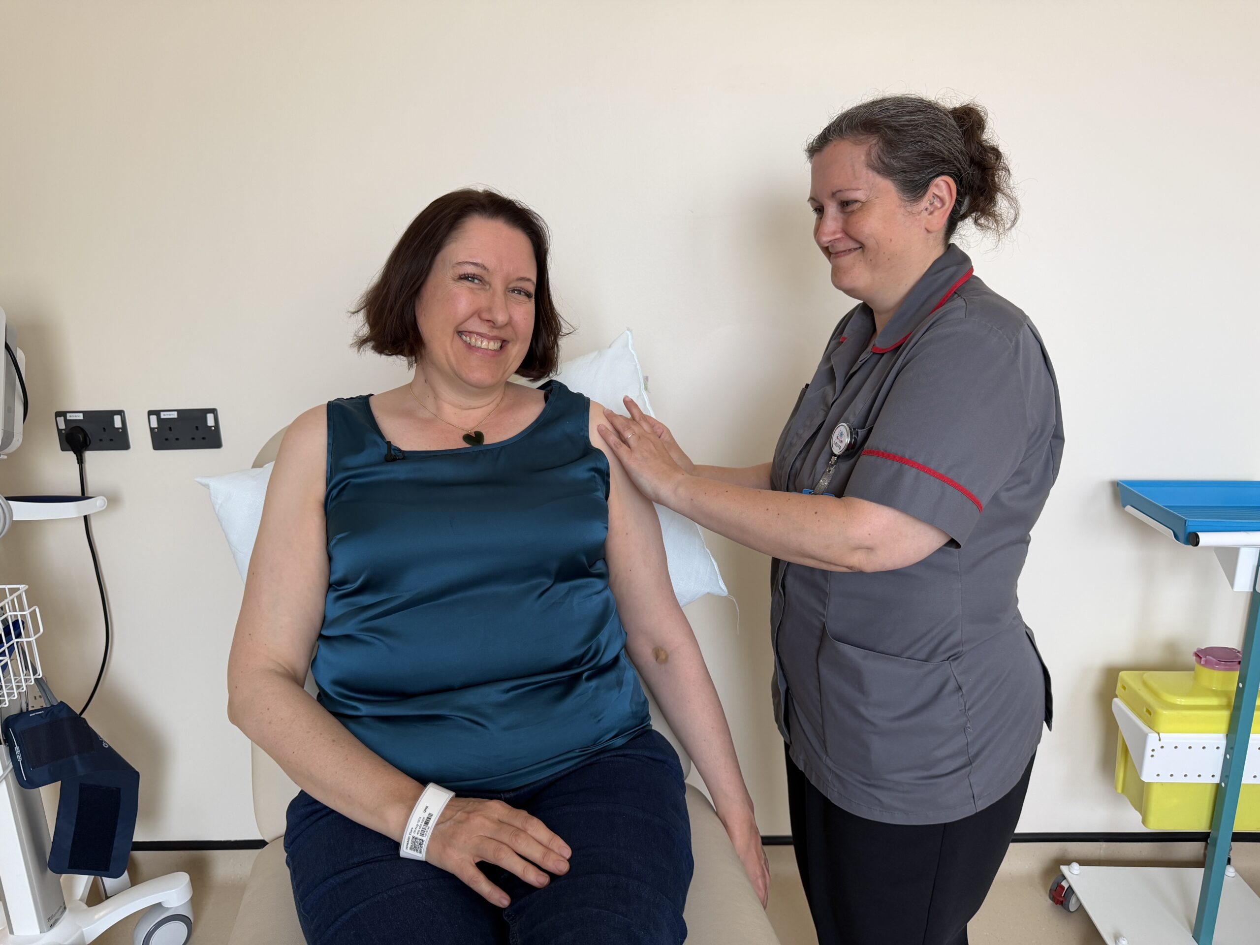 A healthcare professional is smiling and gently touching the shoulder of a seated patient in a medical setting. The patient is also smiling and wearing casual clothing. Medical equipment is visible in the background.